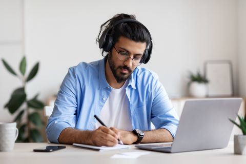 Man sitting at a desk writing in a notebook and wearing headphones